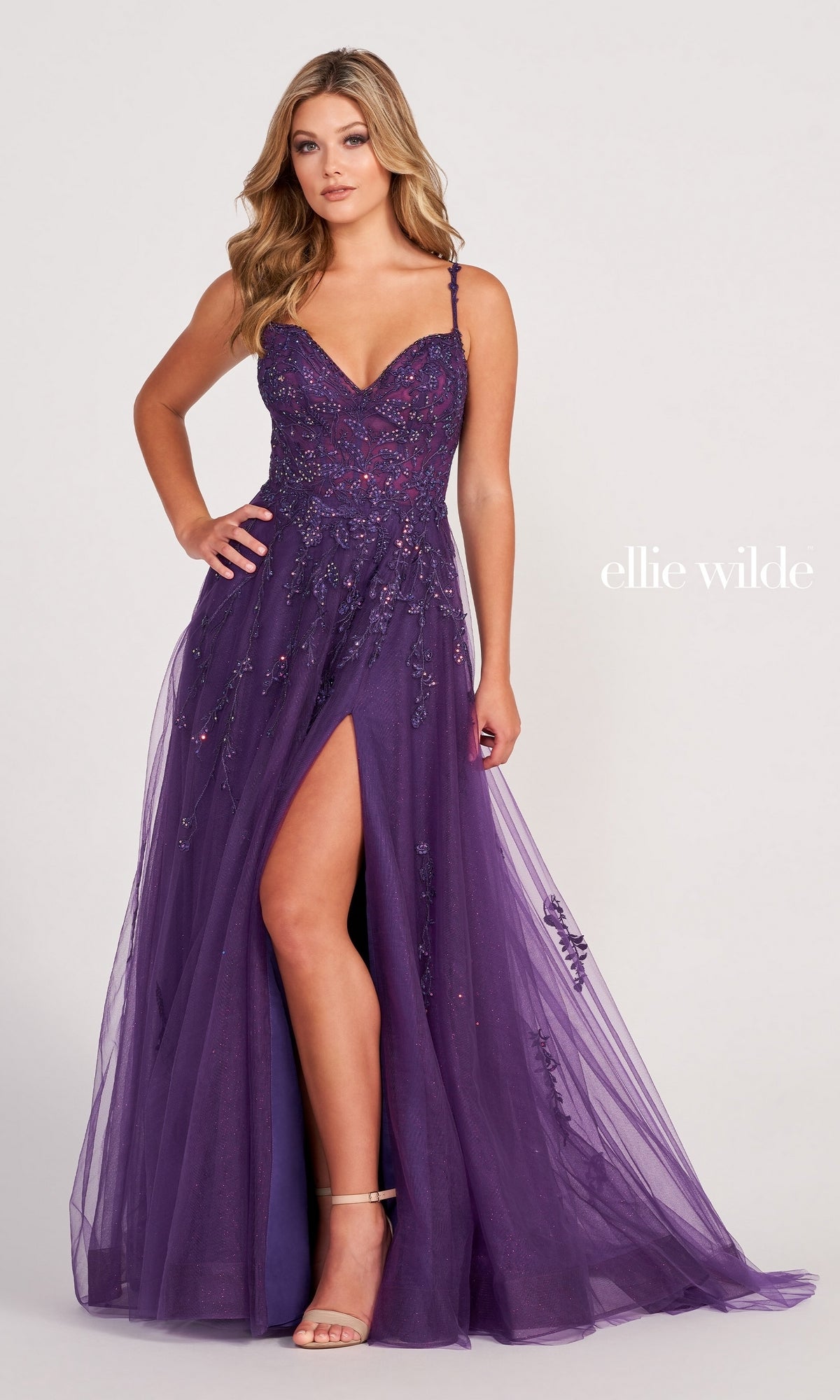 A woman stands against a light background in the Ellie Wilde Long Glitter-Tulle A-Line Formal Dress, featuring a purple hue, spaghetti straps, embellishments, and a thigh-high slit. The text